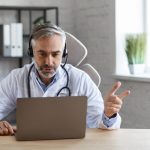 Portrait of senior grey-haired male doctor in his office using laptop for video chat with a patient. Online consultation with doctor for diagnoses and treatment recommendation. Telehealth concept.